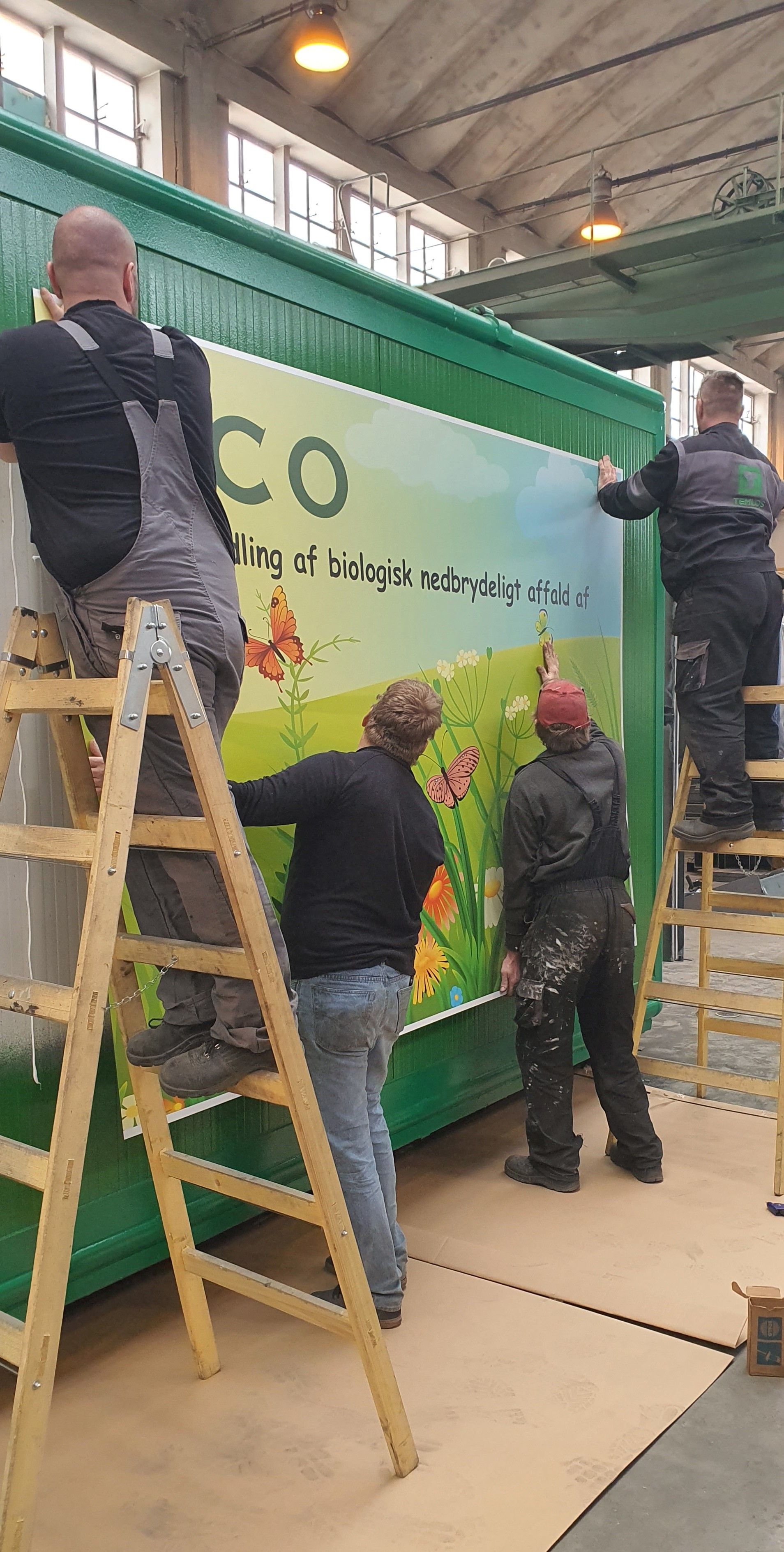 Čtyři pracovníci na štaflích a na zemi pečlivě nalepují velkoplošnou grafiku s logem PERMECO a motivem louky na zelený bok kontejneru.