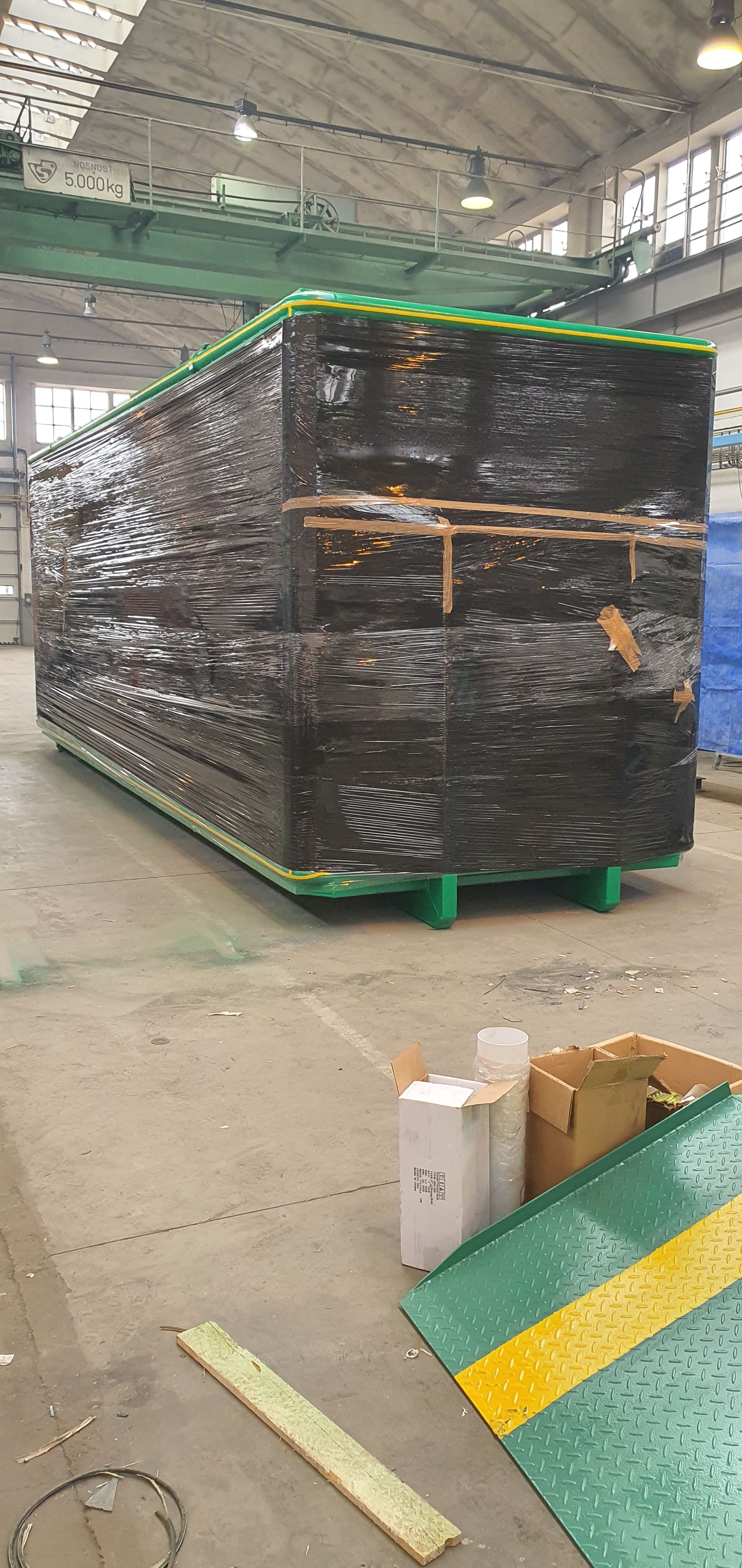 Velký biofermentor kompletně zabalený do černé ochranné fólie, připravený k přepravě ve výrobní hale. V popředí je vidět část zelené nájezdové rampy a kartonové krabice.