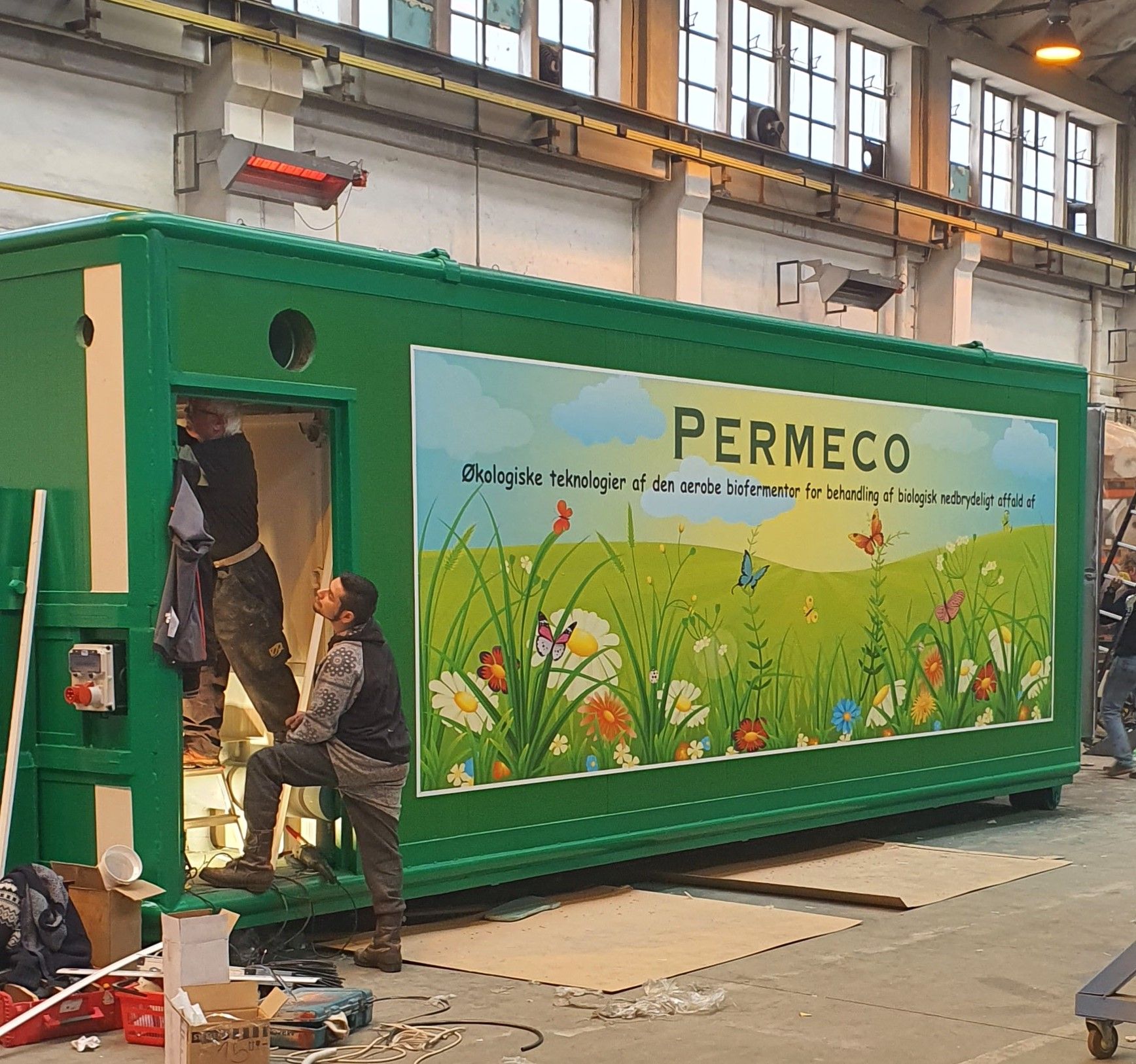 Dělníci v montážní hale pracují na velkém zeleném kontejneru s nápisem PERMECO. Bok kontejneru zdobí grafika rozkvetlé louky a dánský text popisující ekologickou technologii.