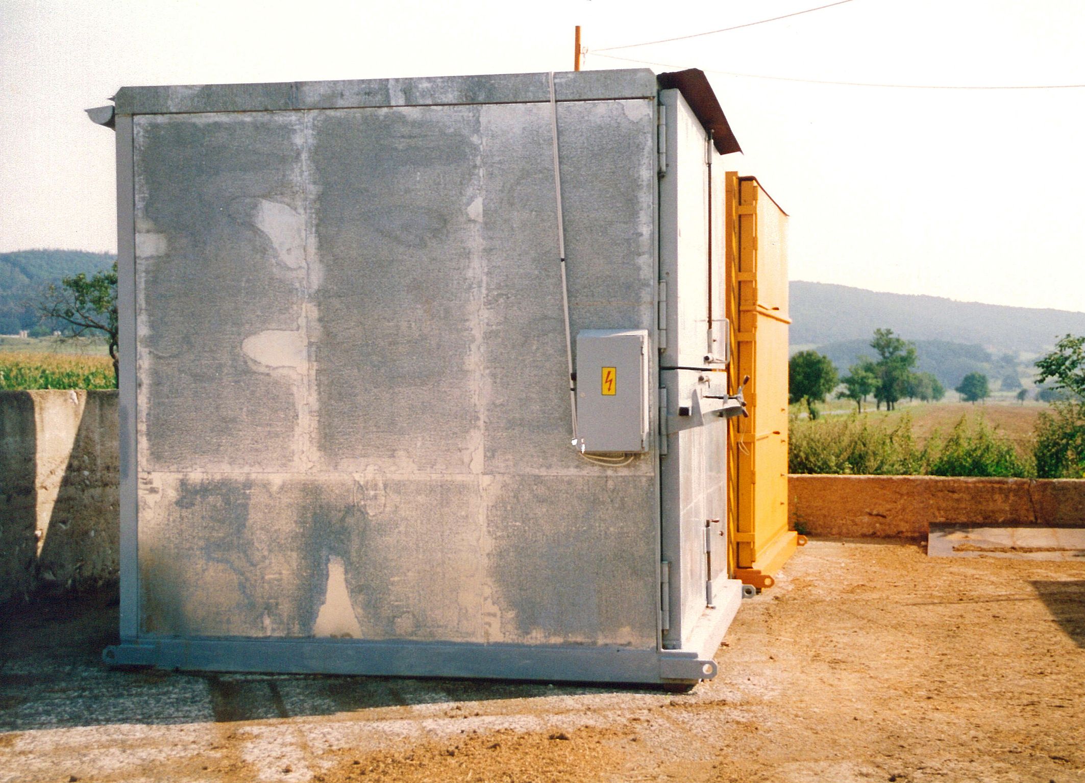 Dobová fotografie staršího typu fermentační jednotky s pozinkovaným pláštěm a elektrickým rozvaděčem na boku, umístěné ve venkovním prostředí u pole.