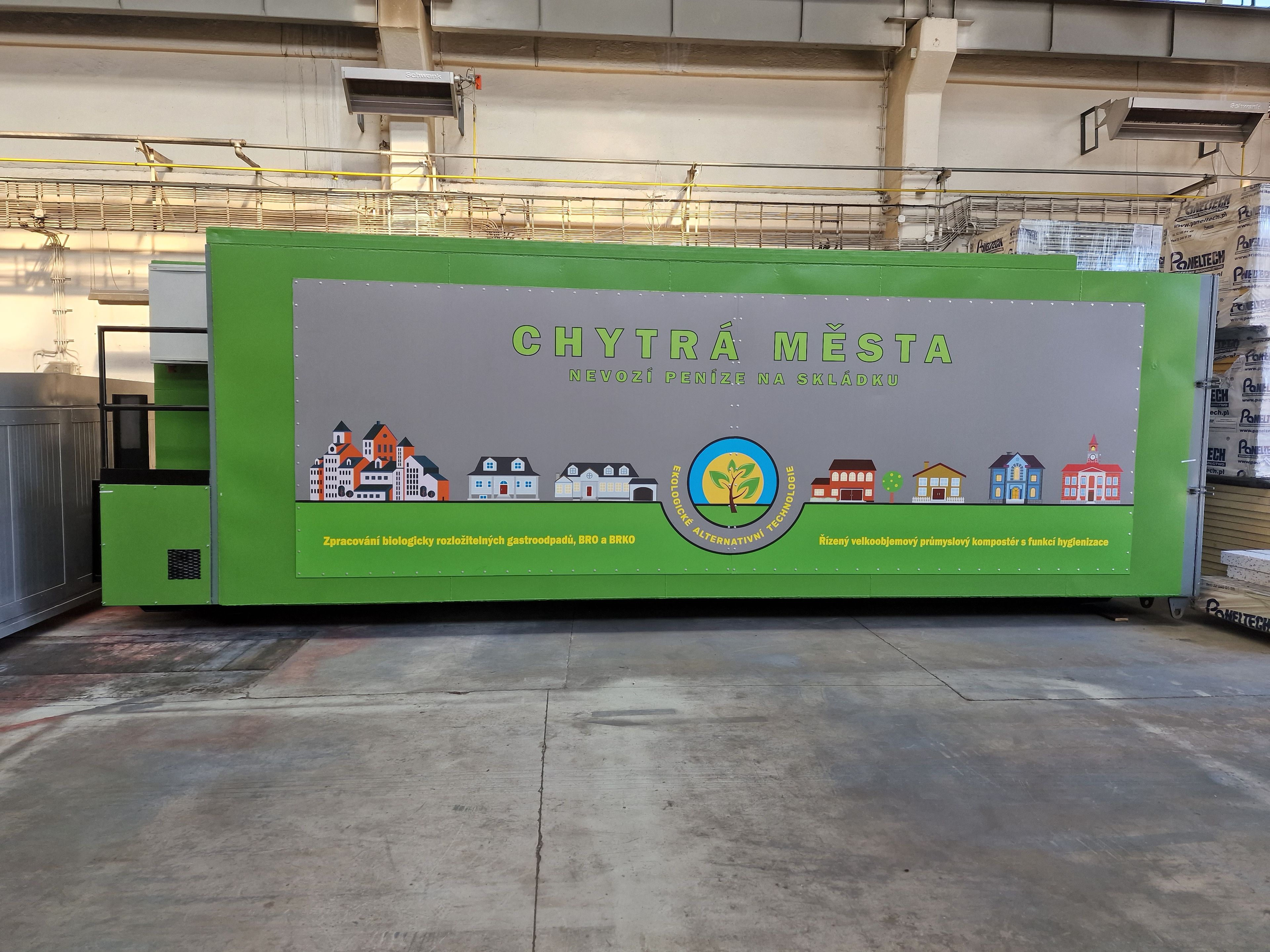Boční pohled na dokončený zelený průmyslový kompostér. Na boku je velký panel s logem, sloganem „Chytrá města nevozí peníze na skládku“ a ilustrací městské zástavby.