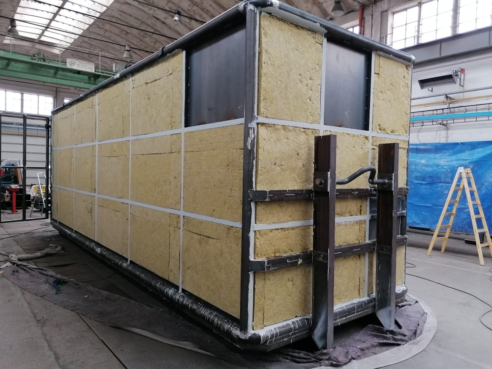 Ocelová konstrukce stěny kontejneru, jejíž rošt je vyplněn deskami žluté minerální vaty pro tepelnou izolaci. Konstrukce stojí na podlaze ve výrobní hale.