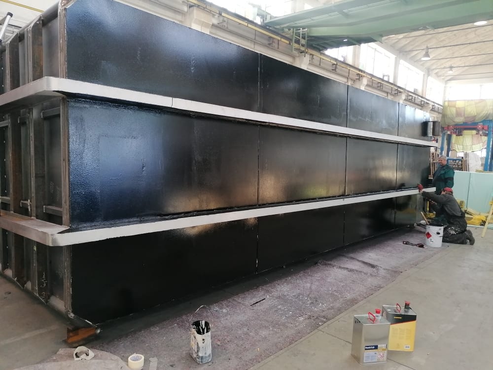 Dva pracovníci v ochranných oblecích natírají velkou ocelovou konstrukci (pravděpodobně vnitřní nádrž) černou barvou. Konstrukce má výrazné horizontální výztuhy.