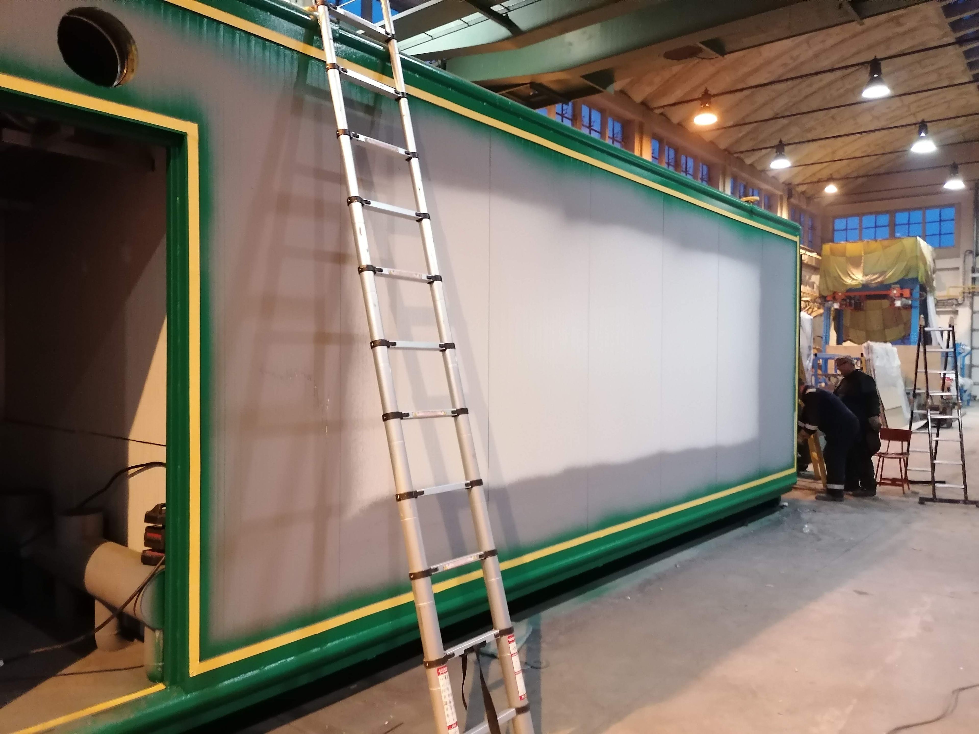 Průmyslová hala, kde probíhá povrchová úprava dlouhého kontejneru. Bok je natřen šedou základovou barvou se zeleným olemováním, v popředí stojí hliníkový žebřík.