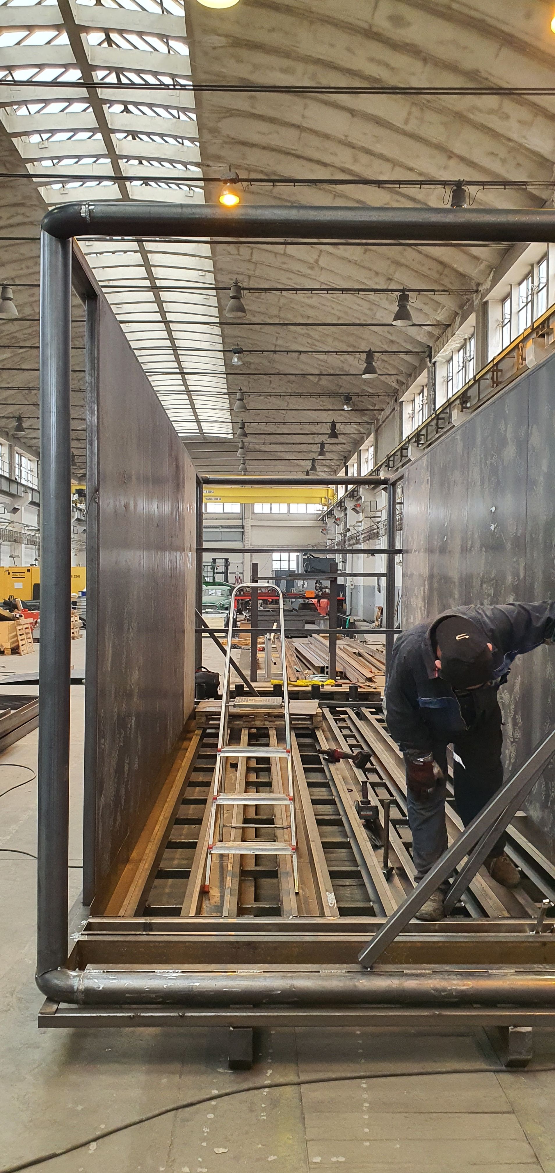 Dělník ve výrobní hale montuje podlahovou část ocelového rámu biofermentoru. Nad ním je vidět otevřená střešní konstrukce a světlíky haly.