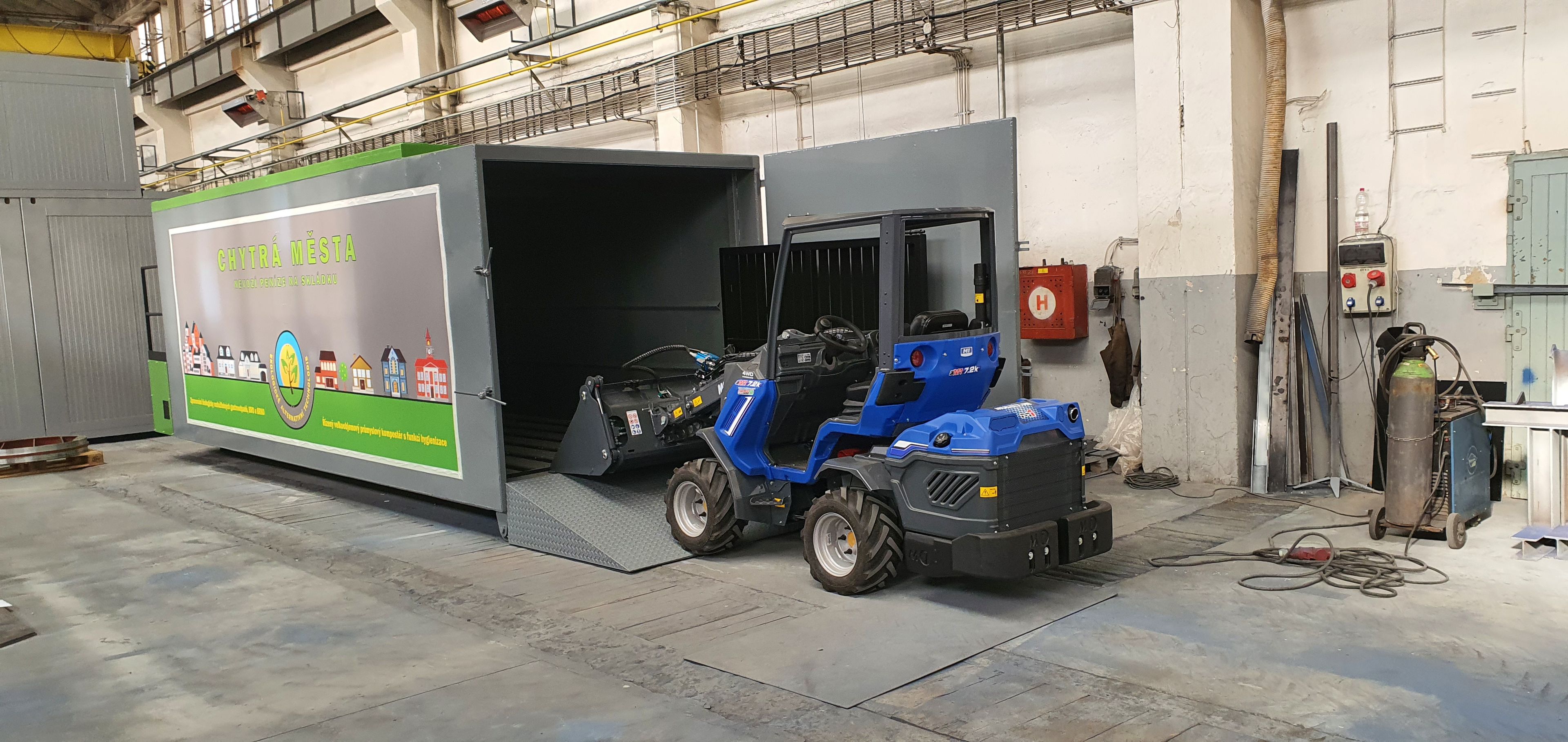 Celkový pohled na zelený biofermentor „Chytrá města“ s otevřenými vraty násypky, ke kterému přijíždí malý modrý kloubový nakladač.