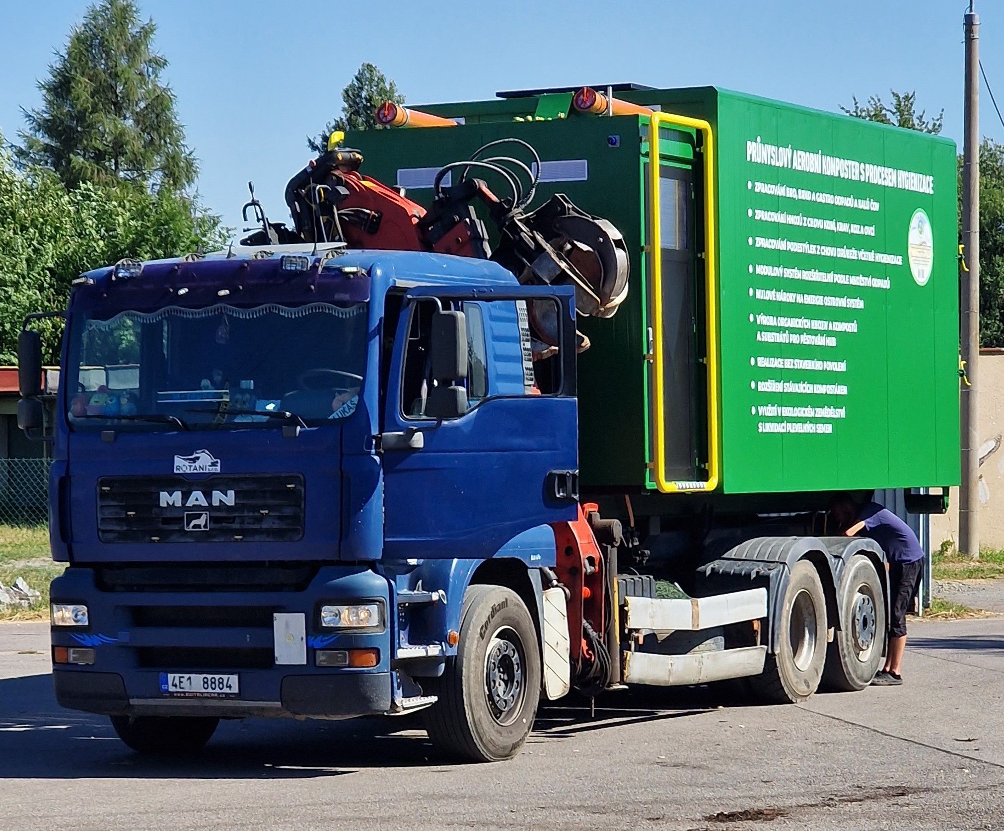 Modrý nákladní automobil s hydraulickou rukou manipuluje se zeleným kontejnerem. Na boku kontejneru je nápis „Průmyslový aerobní kompostér s procesem hygienizace“ a seznam jeho funkcí.