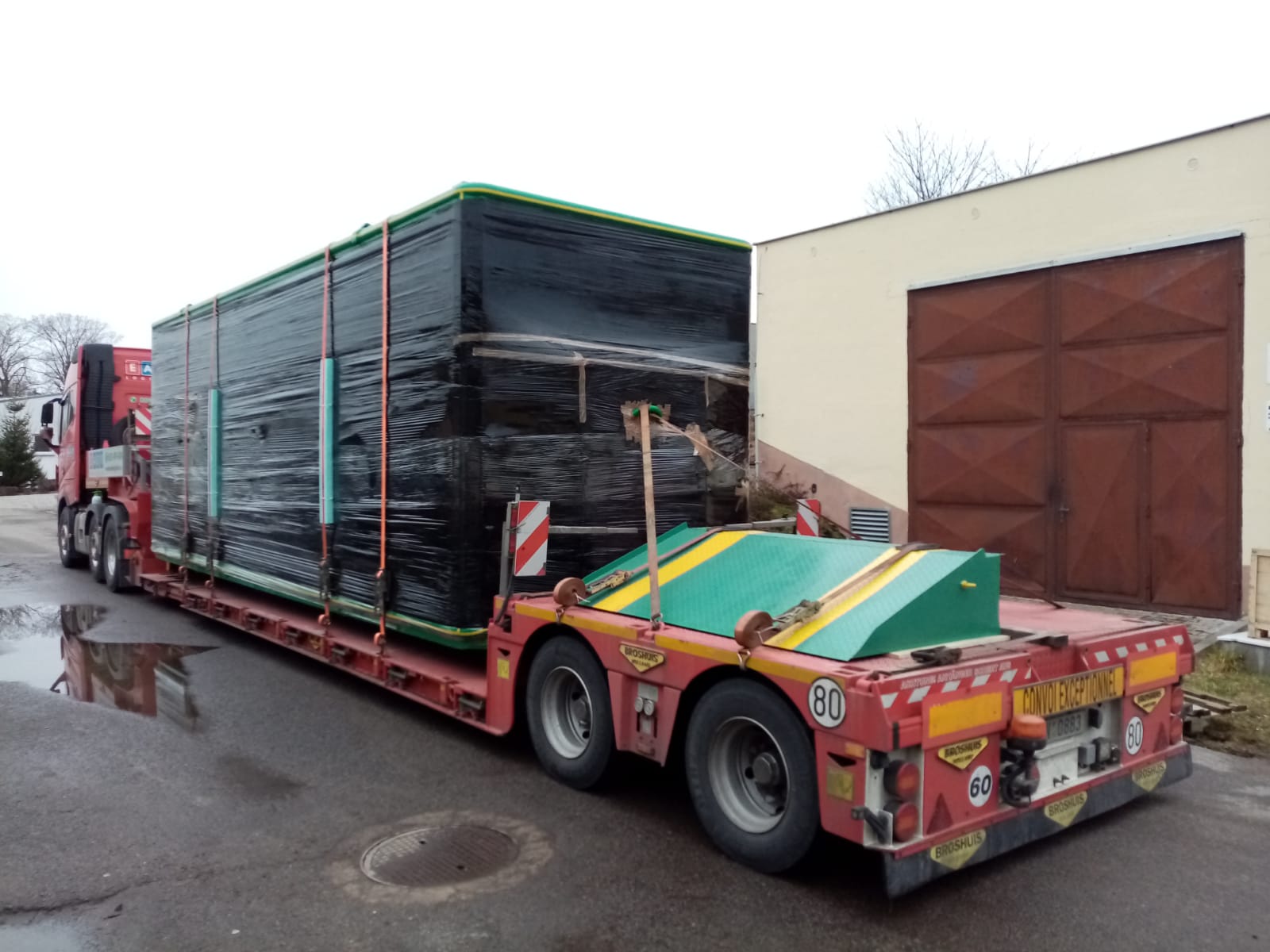 Velký biofermentor zabalený v černé ochranné fólii naložený na podvalníku nákladního automobilu. Náklad je zajištěn popruhy a připraven k transportu v sychravém počasí.