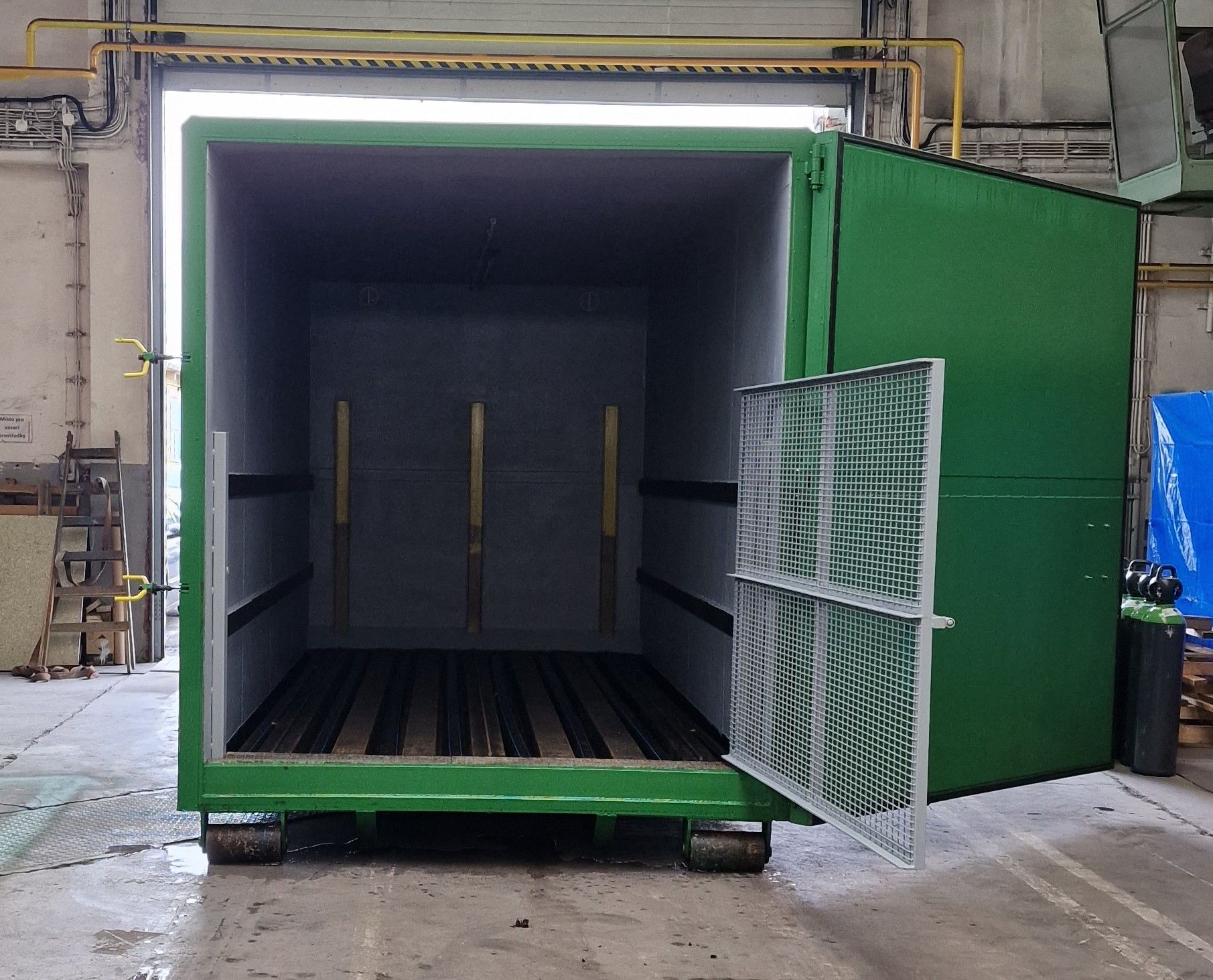 Otevřený zelený biofermentor. Pohled dovnitř ukazuje roštovou podlahu a šedé stěny. Na pravém křídle otevřených vrat je vyklopena vnitřní bezpečnostní mříž.