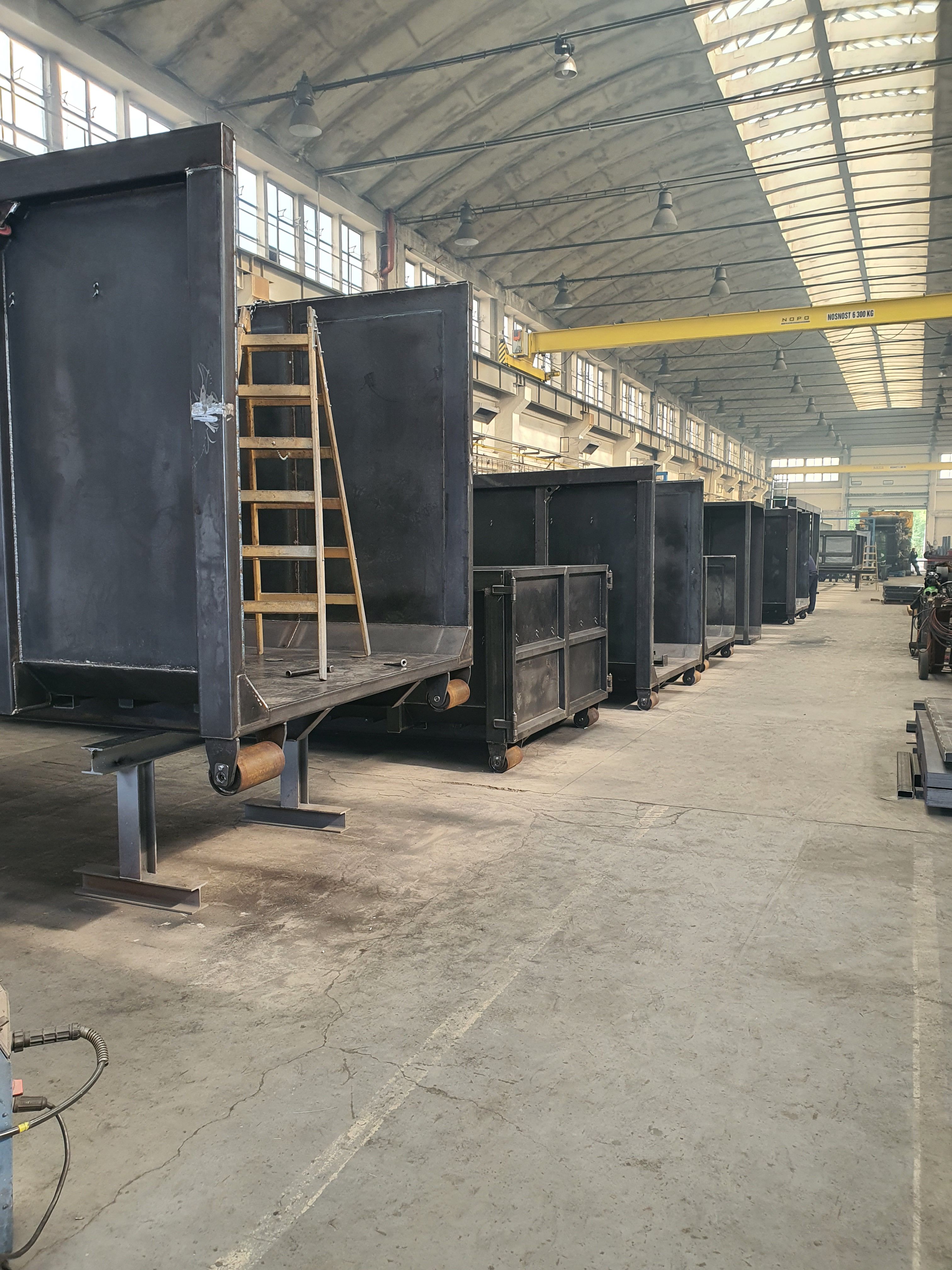Pohled do dlouhé výrobní haly s řadou svařených ocelových koster kontejnerů v surovém stavu. U prvního skeletu je opřený dřevěný žebřík, nad hlavami je žlutý mostový jeřáb.