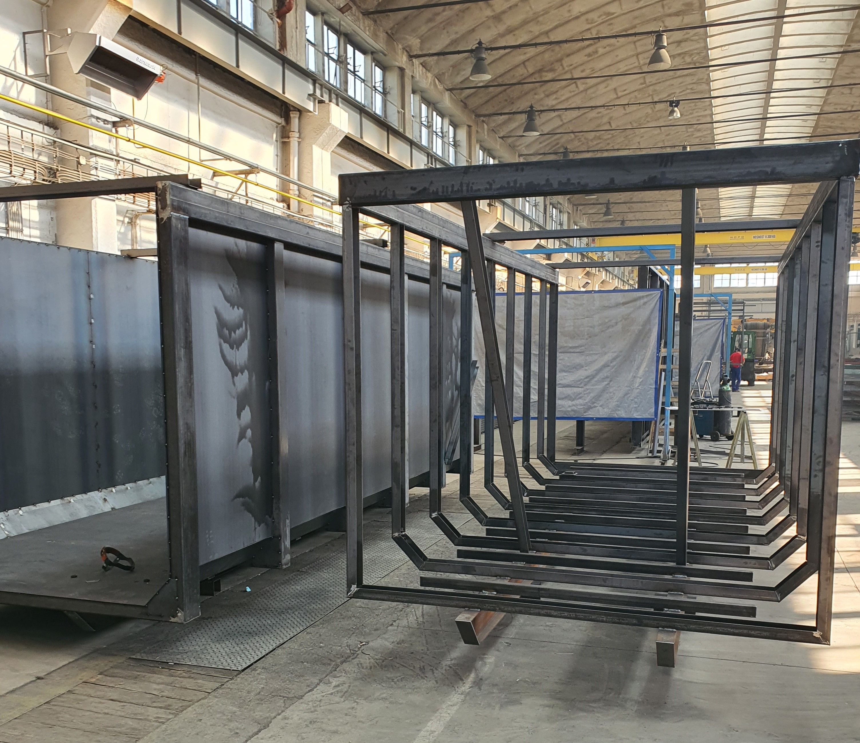 Řada ocelových rámů (koster) pro biofermentory seřazená ve výrobní hale. Konstrukce jsou zatím bez opláštění a čekají na další fázi výroby.