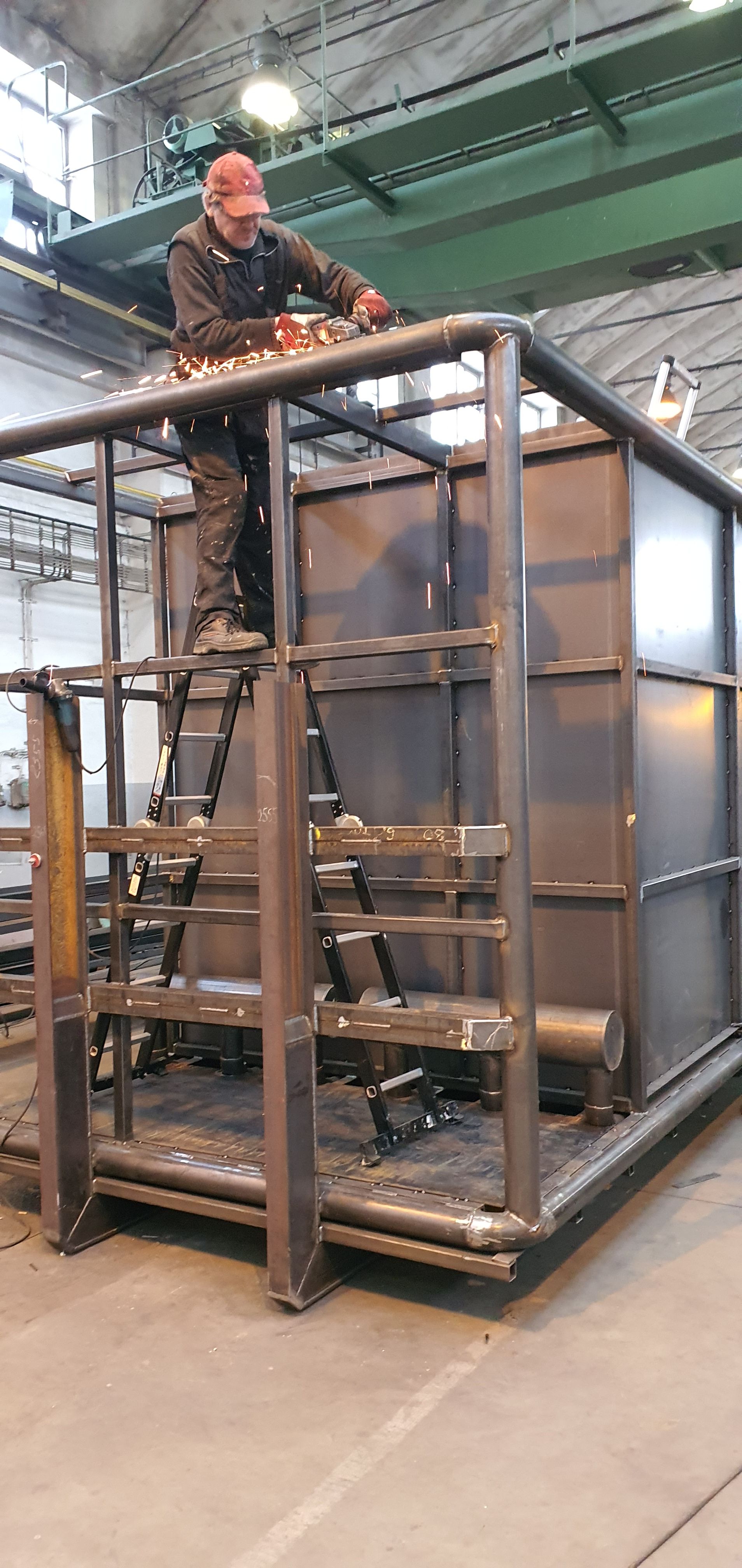 Dělník stojící na žebříku brousí horní část ocelového rámu konstrukce, přičemž odletují jiskry. Jedná se o hrubou výrobu skeletu zařízení.